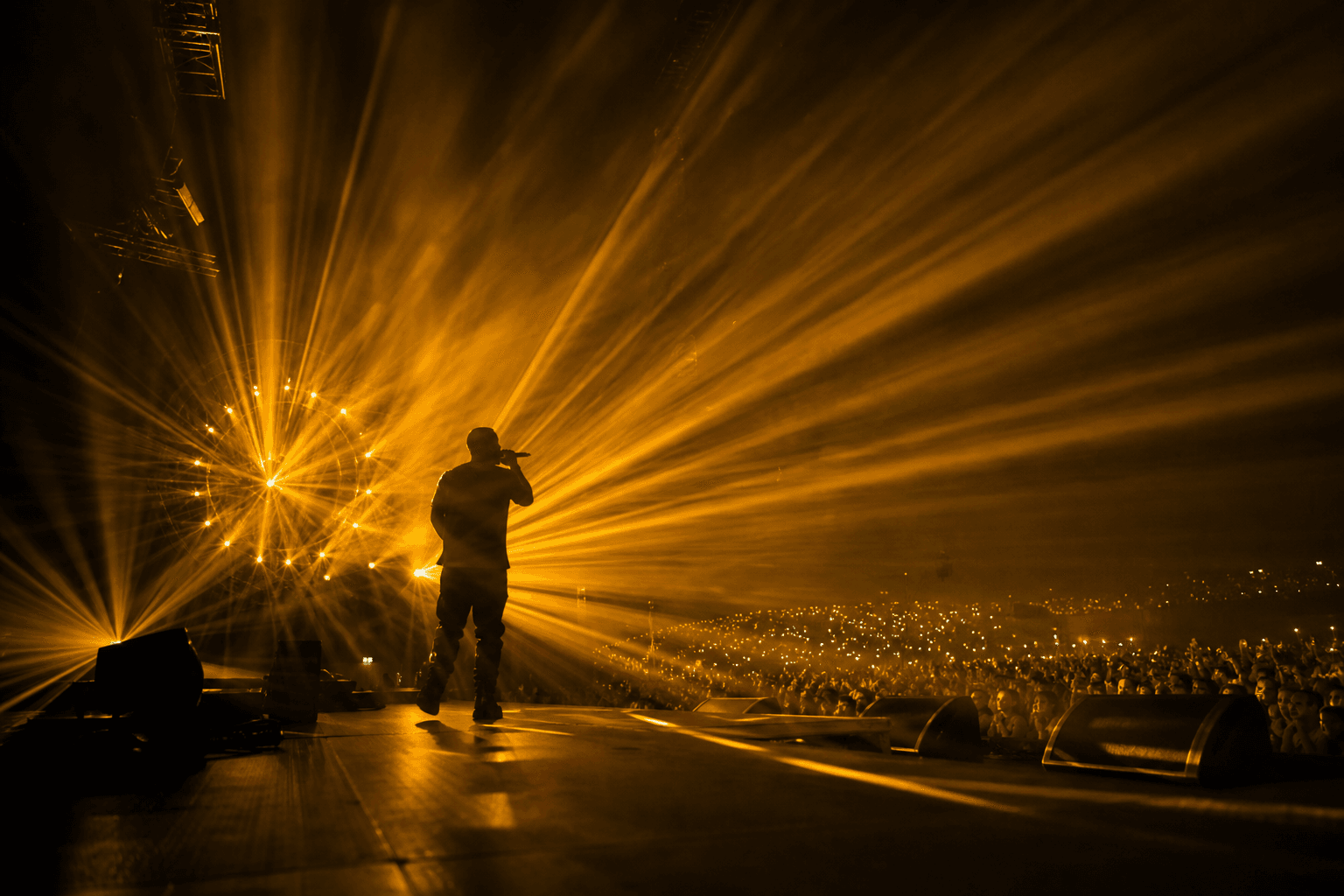 Performer on a concert stage with amber light beams made visible by haze above a large crowd